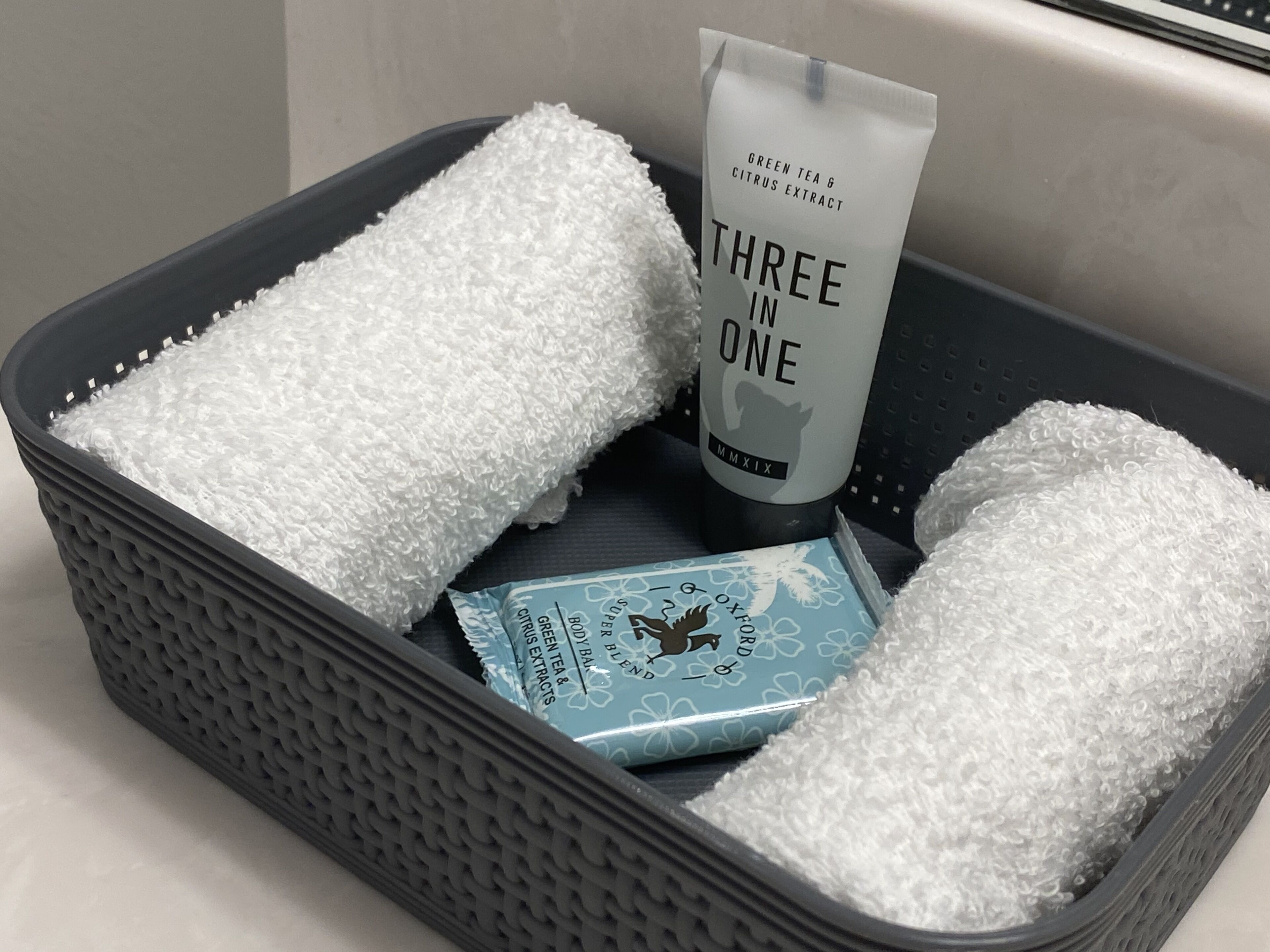 Un panier gris texturé contenant deux serviettes blanches, un tube d’extrait de thé vert et d’agrumes, et une boîte bleue ornée de motifs floraux.