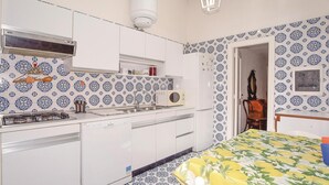 Uma cozinha compacta com armários brancos, fogão a gás, micro-ondas e pia. O chão tem ladrilhos estampados e as paredes, azulejos decorativos em azul e branco.