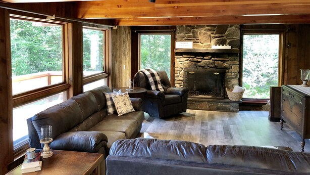 A cozy cabin interior with a stone fireplace, leather sofas, and large windows offering a view of the forest.
