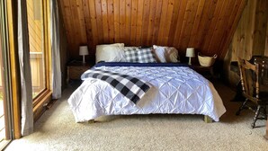 A bedroom with a wooden ceiling, a bed with a white and blue quilt, two bedside tables with lamps, and a chair.
