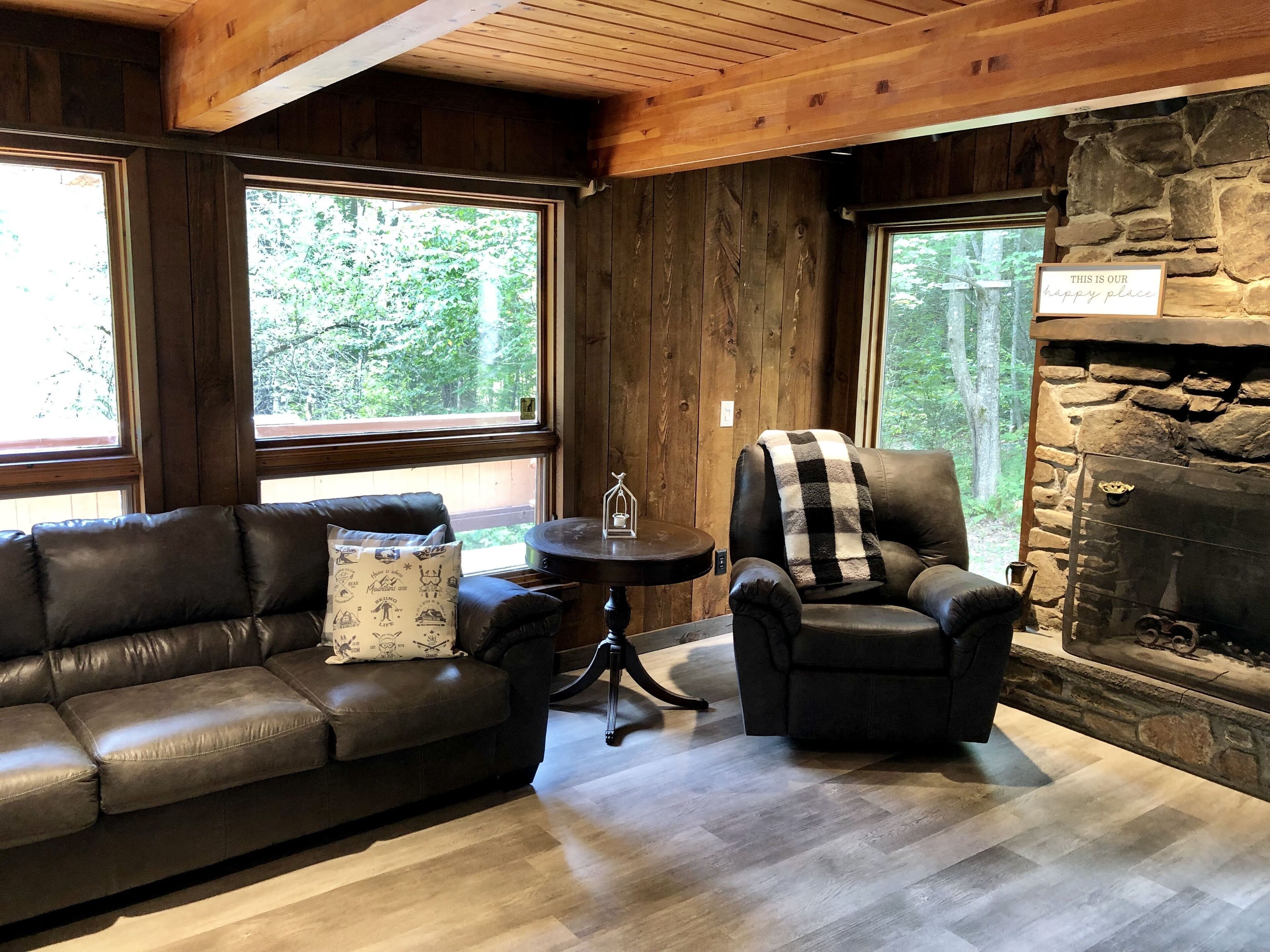 Ein gemütliches Wohnzimmer mit Kamin aus Stein, Ledersofas und großen Fenstern mit Blick auf den Wald.