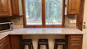 A kitchen with wooden cabinets, a granite countertop, a microwave, three black stools, and a window with a forest view.
