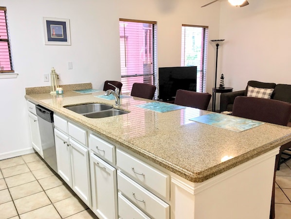 A kitchen with a granite countertop, stainless steel sink, and white cabinetry. There are two windows with blinds, a small framed picture on the wall, and a floor lamp.