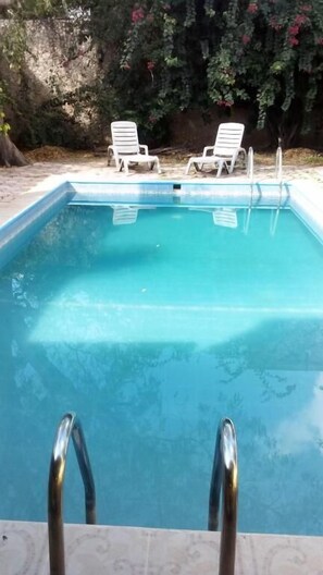 A rectangular swimming pool with clear blue water, surrounded by a tiled deck. There are two white lounge chairs with metal frames and a metal ladder leading into the pool. - Yucatan Vista Inn (Mérida)