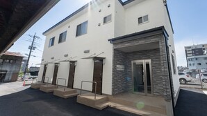Un edificio de dos pisos con un diseño moderno, que presenta un techo plano y una combinación de muros de ladrillo y paredes exteriores blancas. Se observan varias puertas y ventanas, y una pequeña zona de entrada con escalones que conducen a la entrada principal.