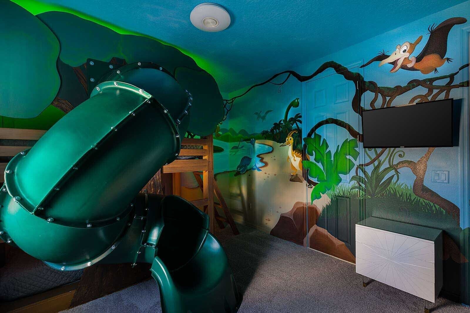 A room with a green slide, a TV on the wall, and jungle-themed wall art.