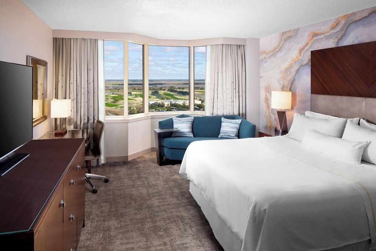Ein Hotelzimmer mit einem großen Bett, einem Schreibtisch, einem Fernseher und Blick auf die Landschaft.