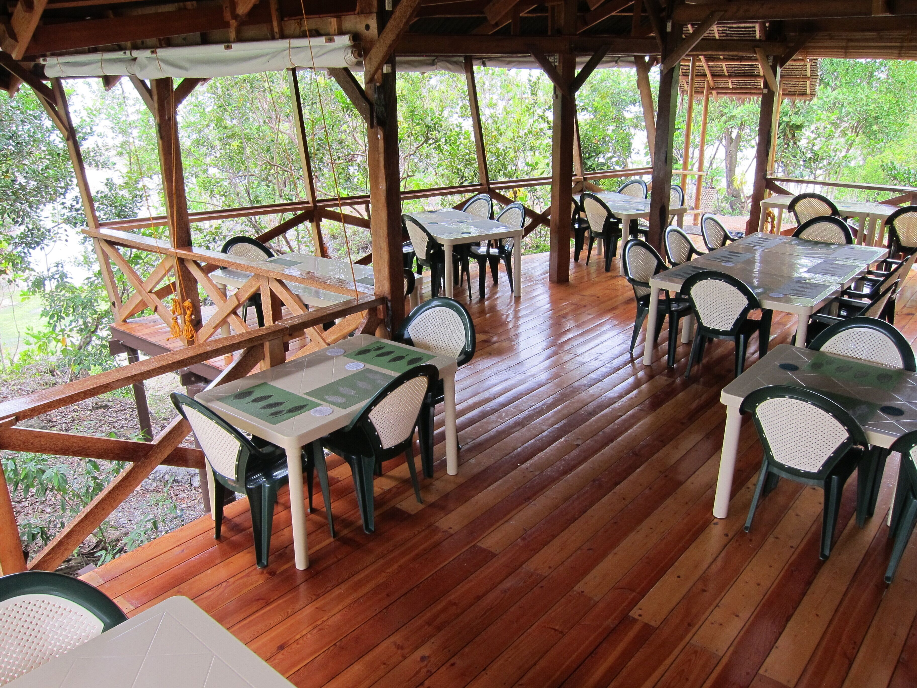An outdoor dining area with wooden tables and chairs, surrounded by a wooden deck and large windows overlooking a forested area.