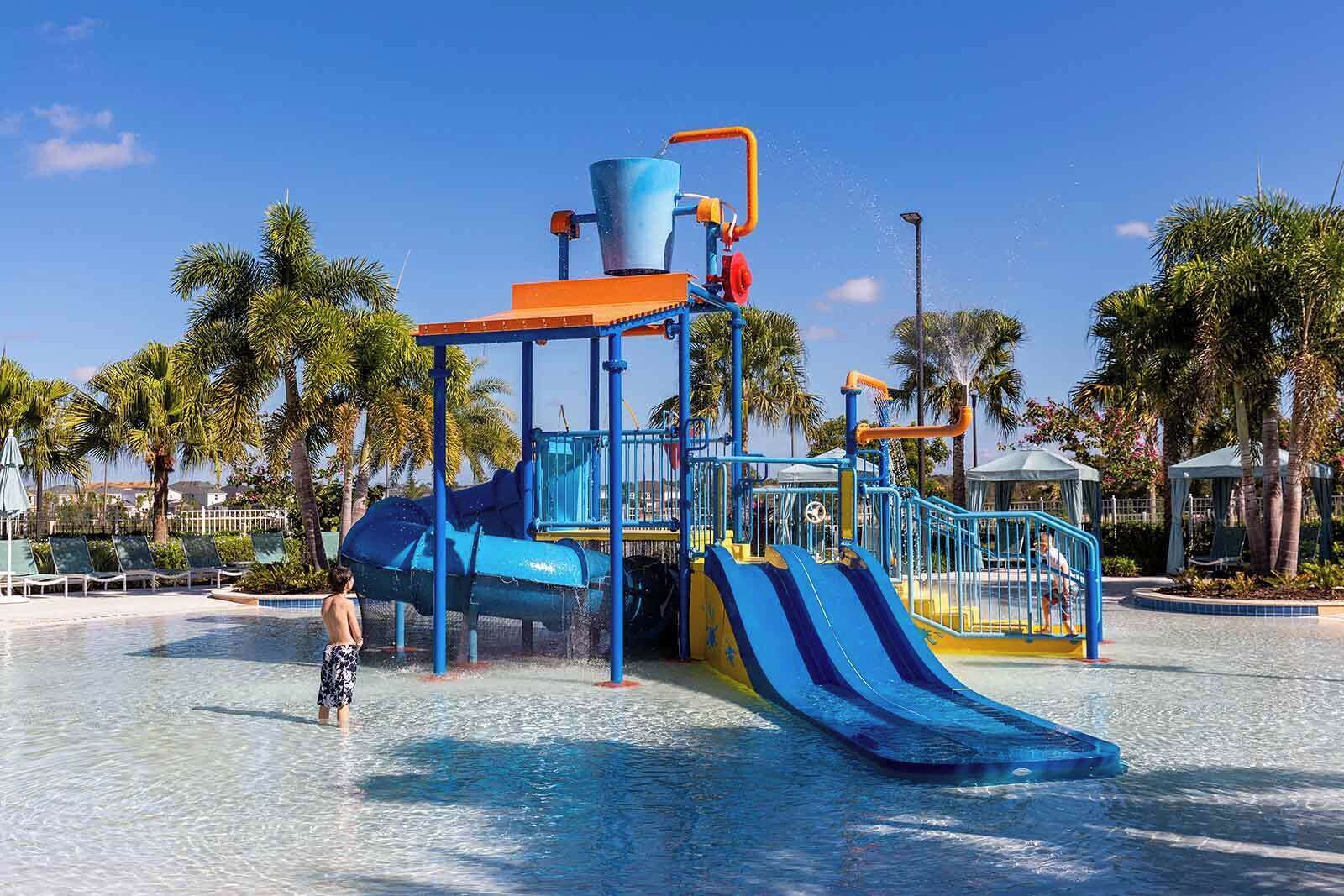 Zona de juegos acuáticos con toboganes y una fuente, rodeada de palmeras y una piscina.