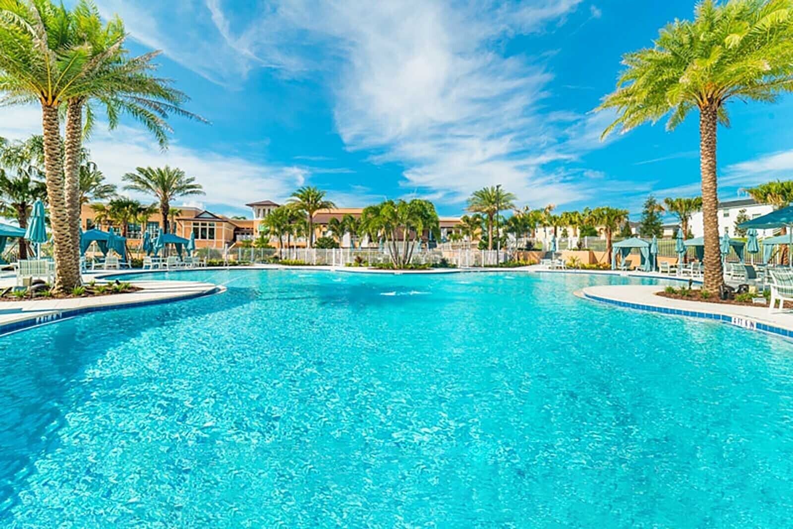 A large, clear swimming pool with lounge chairs and umbrellas, surrounded by palm trees and buildings.