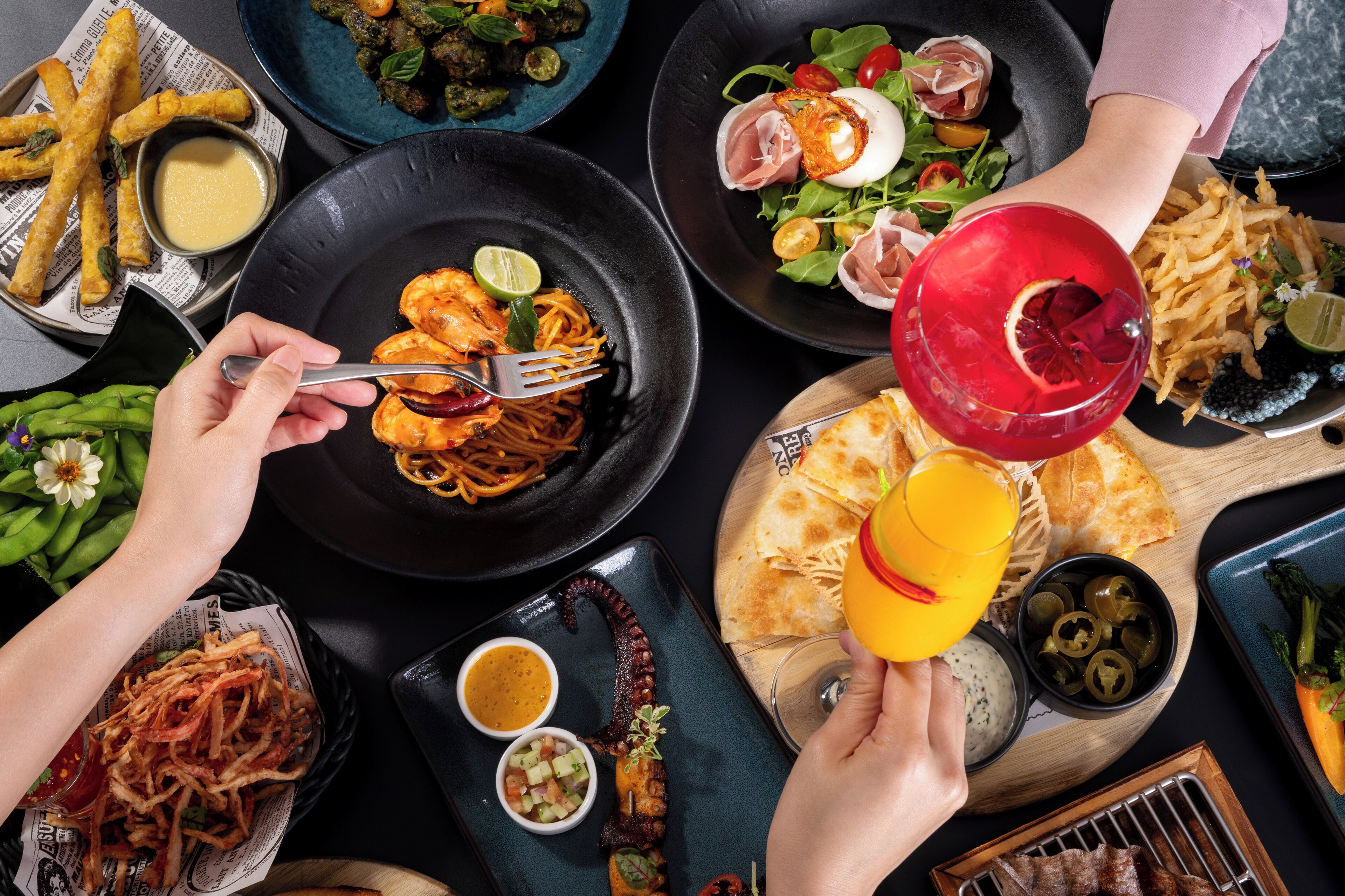 A variety of dishes including shrimp, fries, and salads, with a person holding a drink.