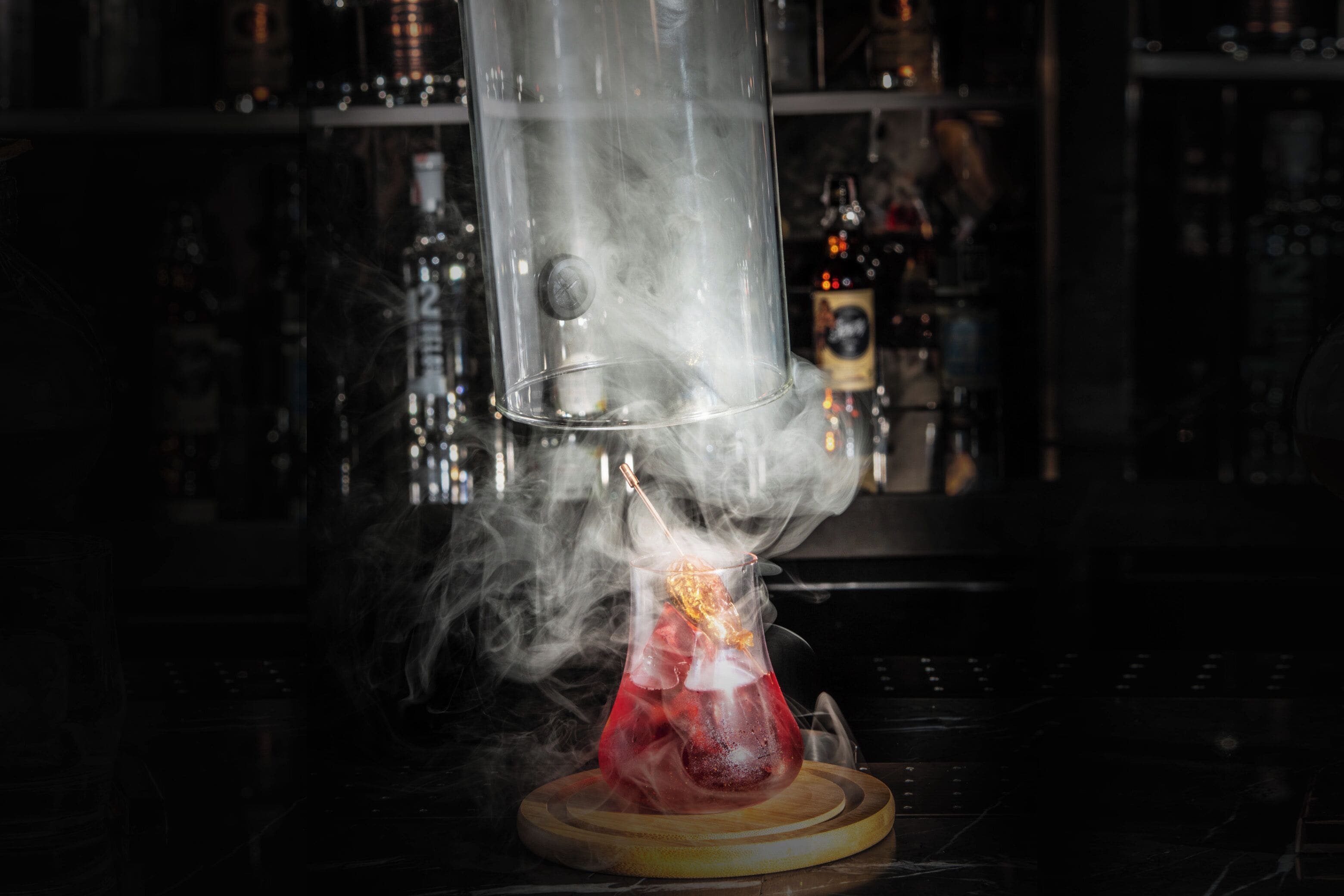 A bar setting with a clear glass container emitting smoke, a cocktail glass on a wooden coaster, and a bar counter with bottles in the background.