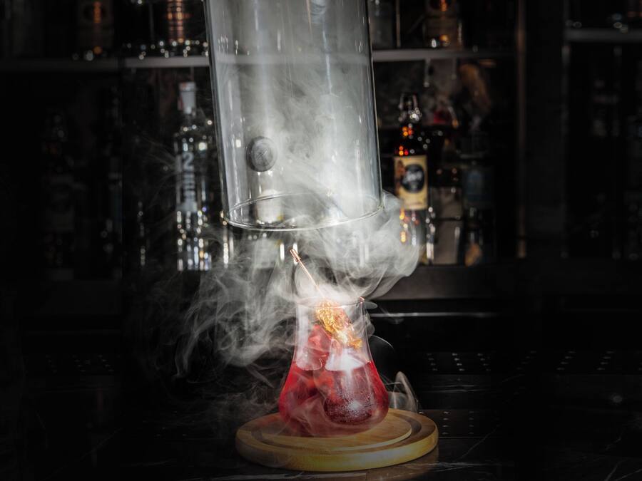 Un ambiente de bar con un recipiente de vidrio transparente que emite humo, una copa de cóctel sobre un posavasos de madera y una barra con botellas al fondo.