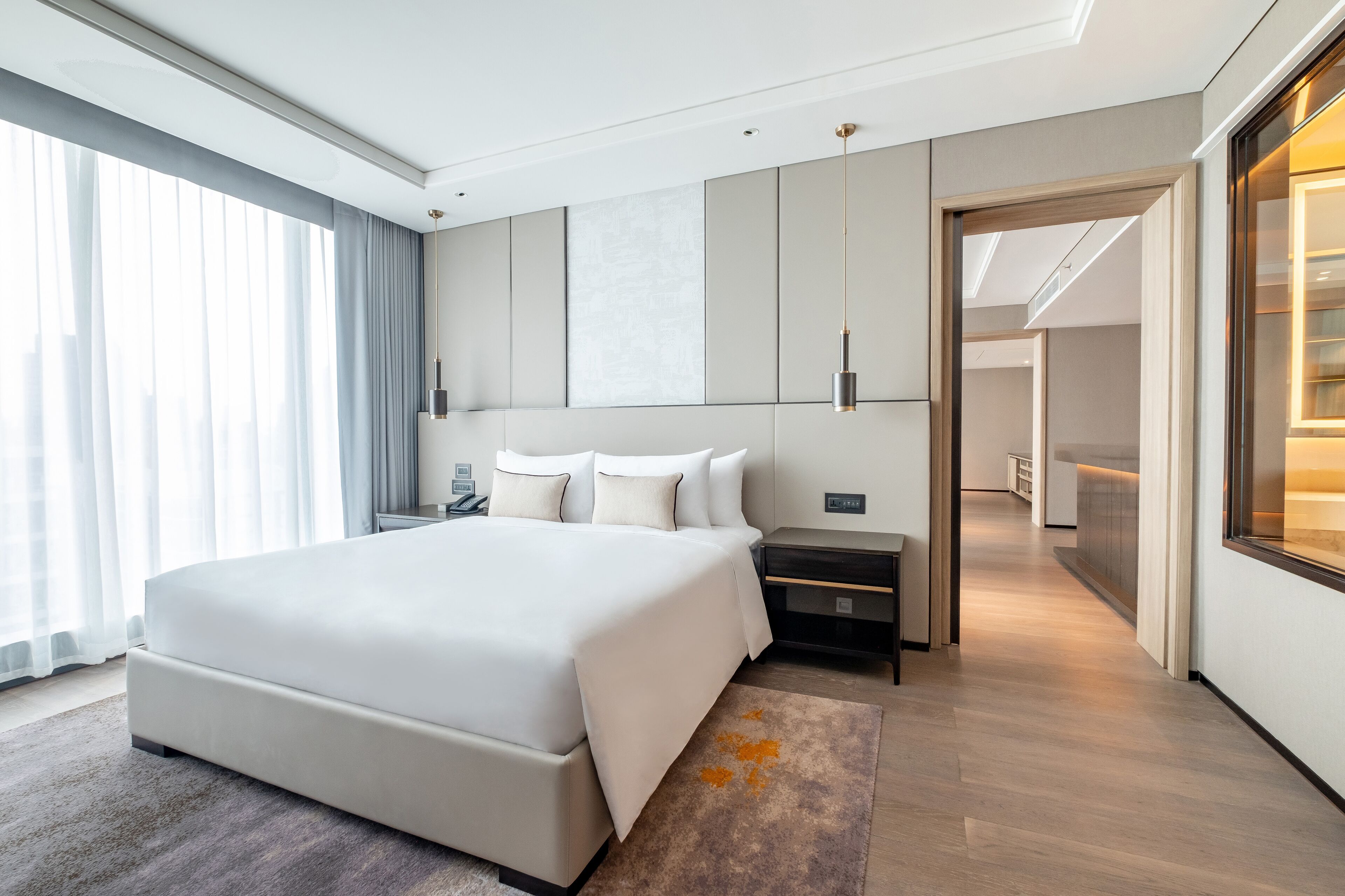 Una habitación de hotel moderna con una cama grande, mesitas de noche, un armario y vistas de la ciudad a través de amplios ventanales.