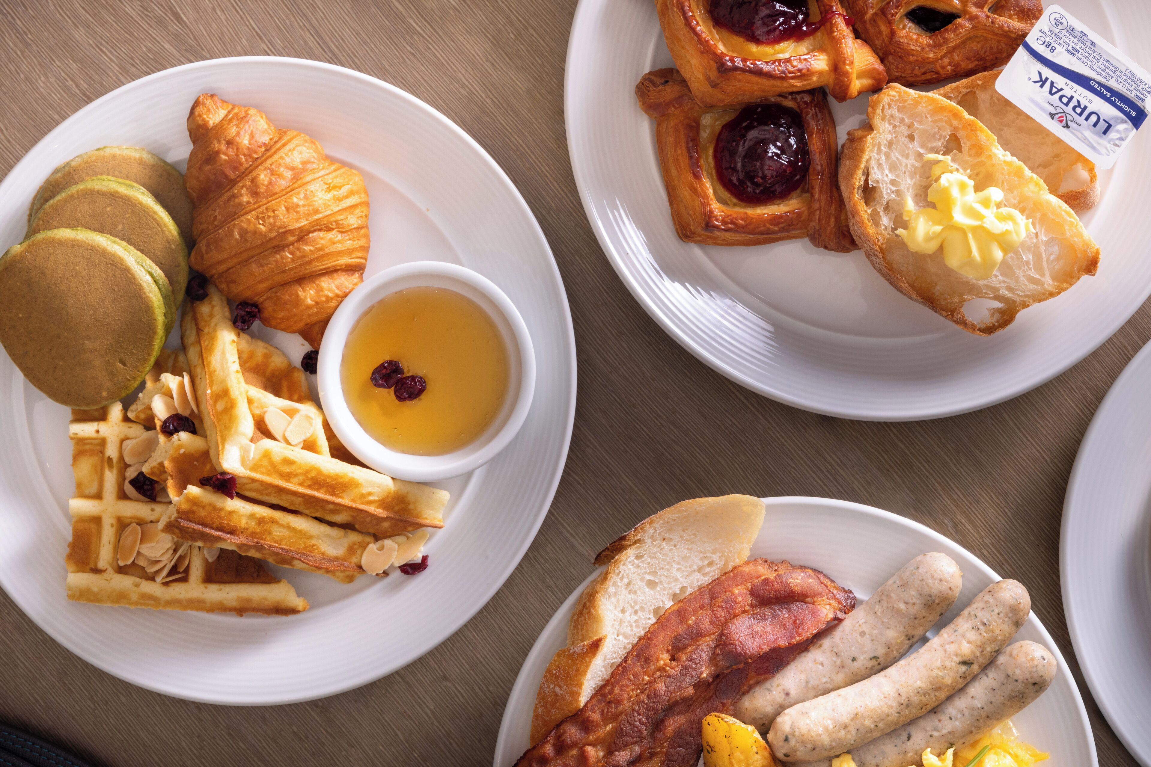 Trois assiettes de nourriture pour le petit-déjeuner, comprenant des crêpes, des croissants et des saucisses.