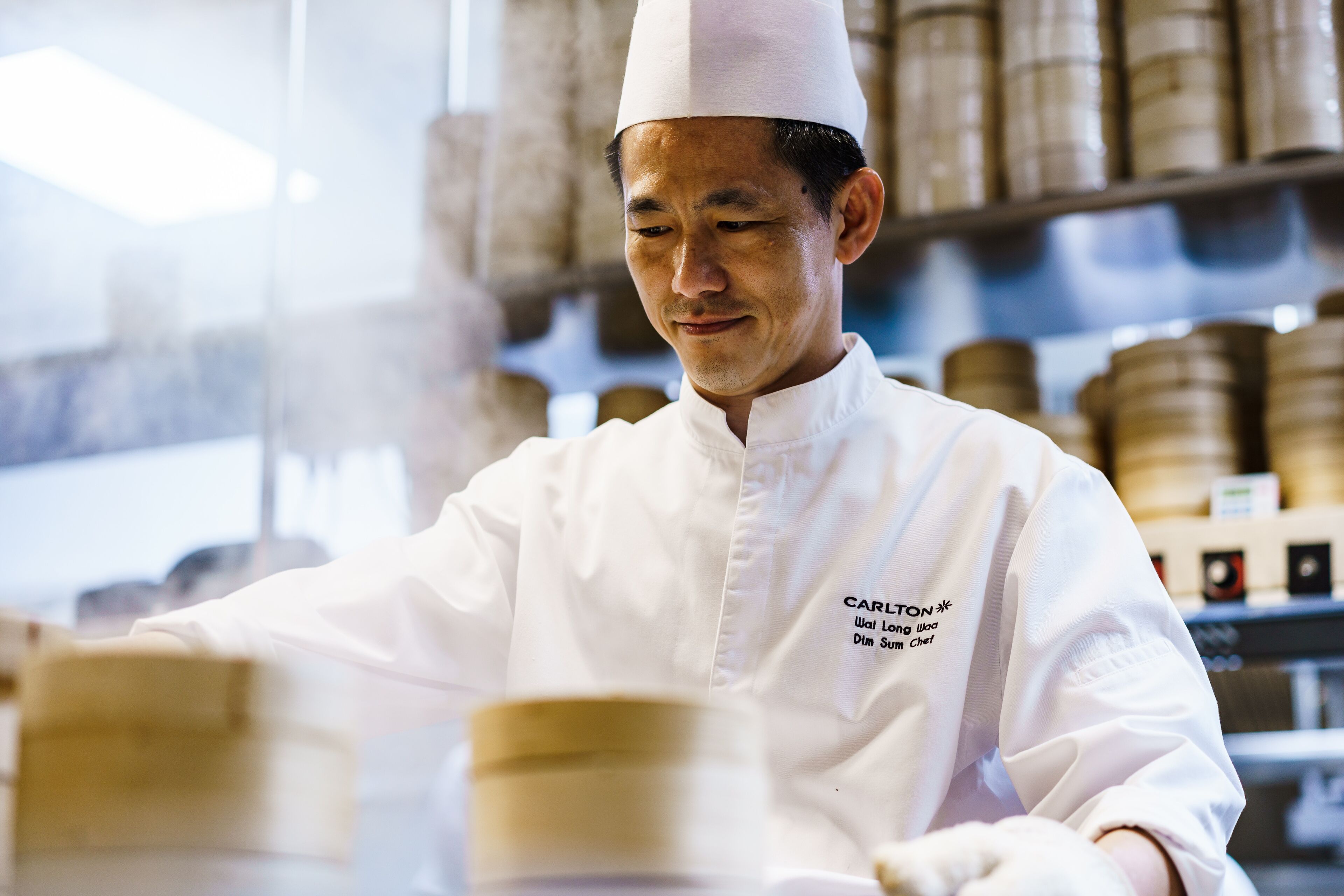 A chef in a white uniform with a name tag, working in a kitchen with shelves of ingredients in the background.