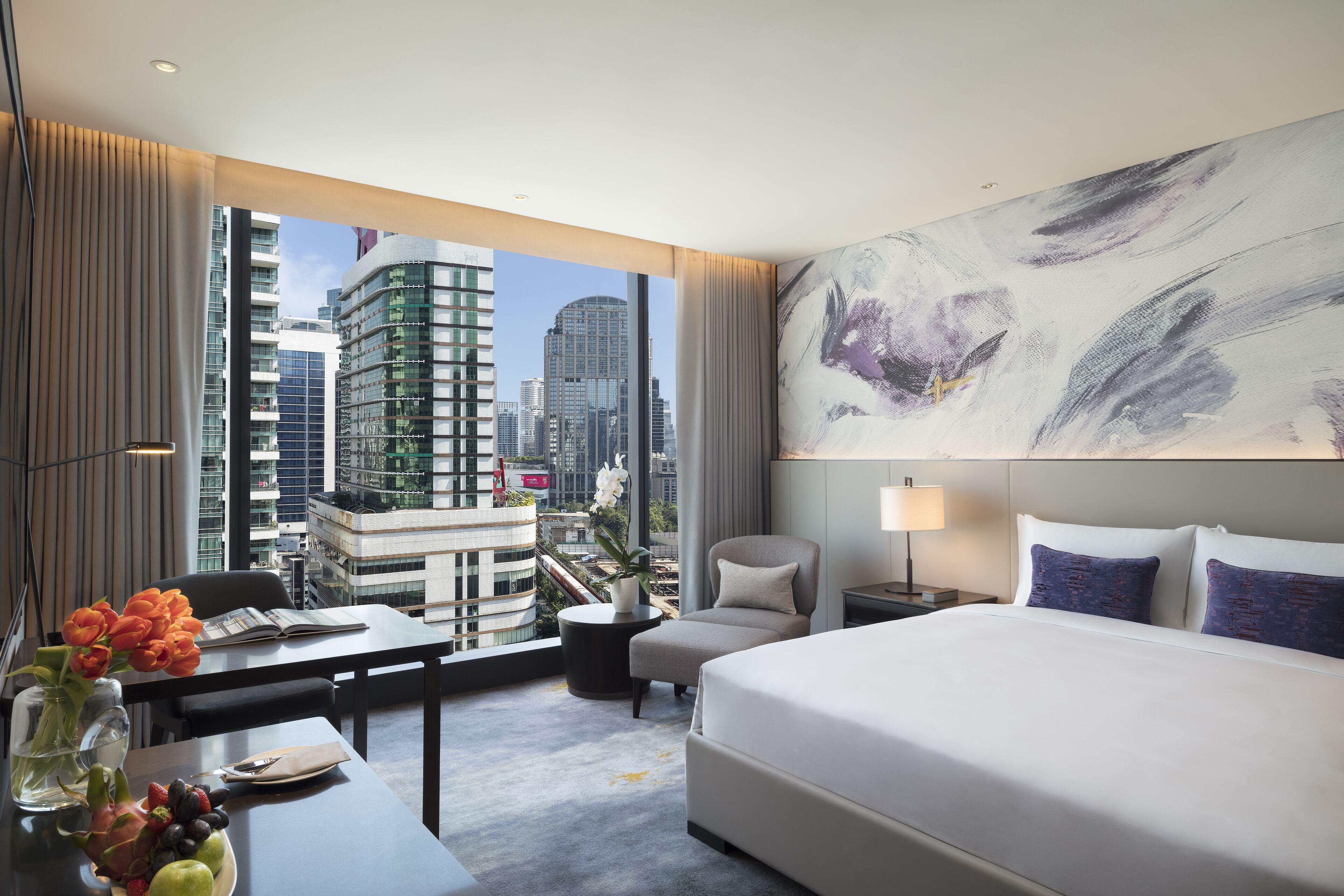 A modern hotel room with a large bed, a desk with a vase of flowers, a chair, and a view of the city skyline through large windows.