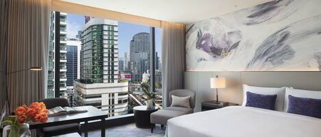 A modern hotel room with a large bed, a desk with a vase of flowers, a chair, and a view of the city skyline through large windows.