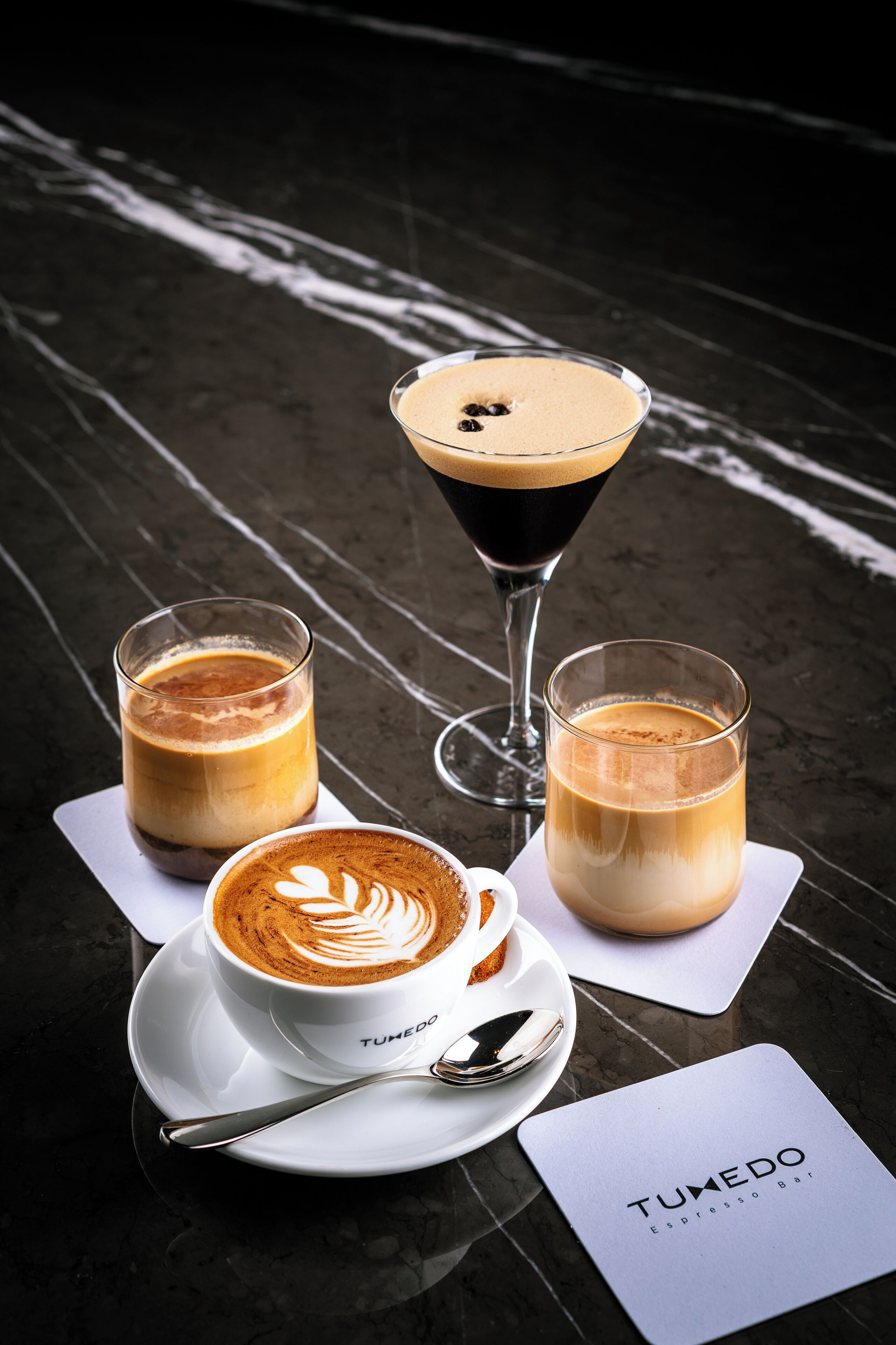Three coffee drinks on a marble table with a menu card labeled "TumedO Espresso Bar".