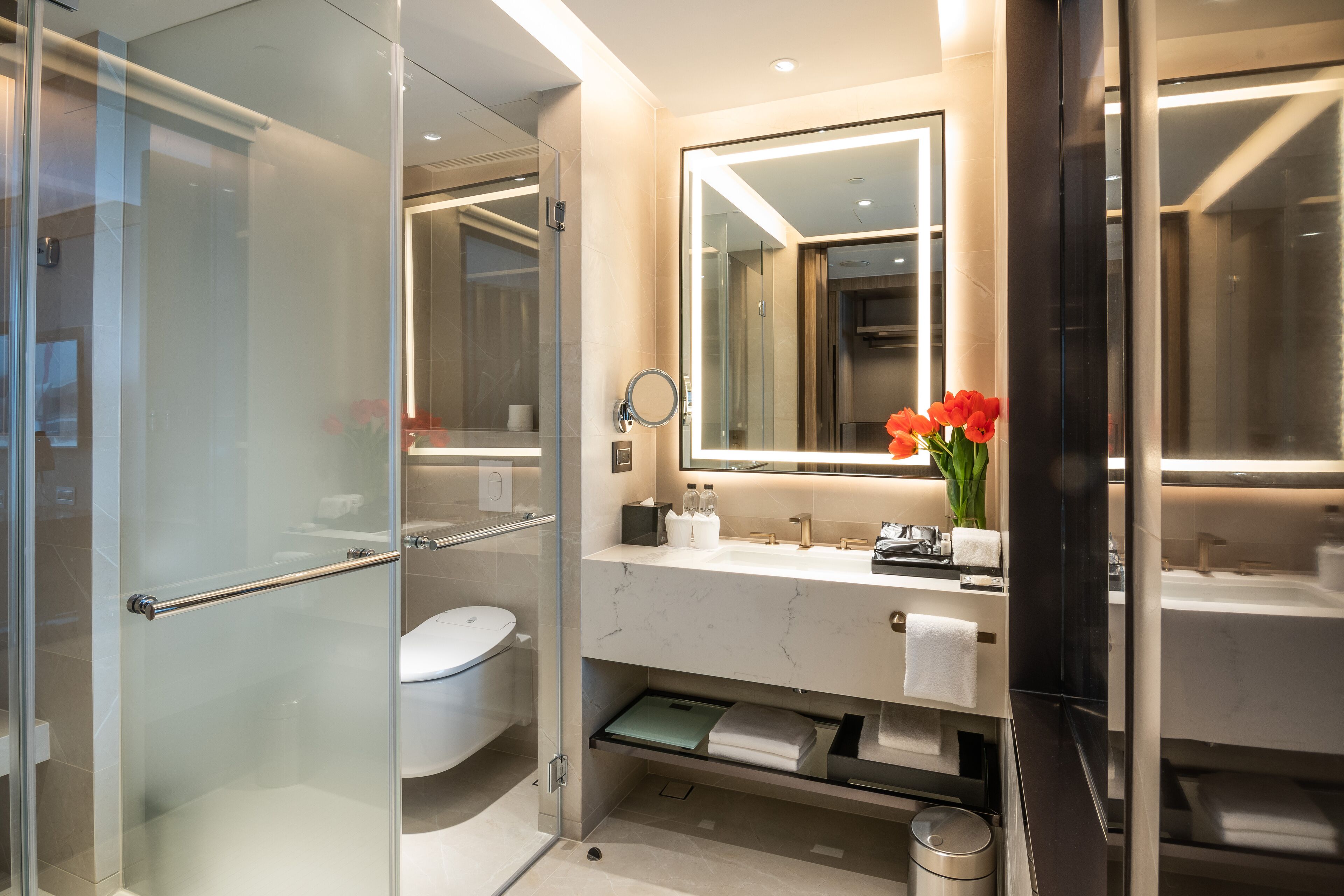 Un baño moderno con cabina de ducha de vidrio, inodoro, tocador con espejo y un florero con flores.