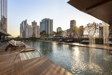 A rooftop pool with lounge chairs, a wooden deck, and a city skyline in the background.
