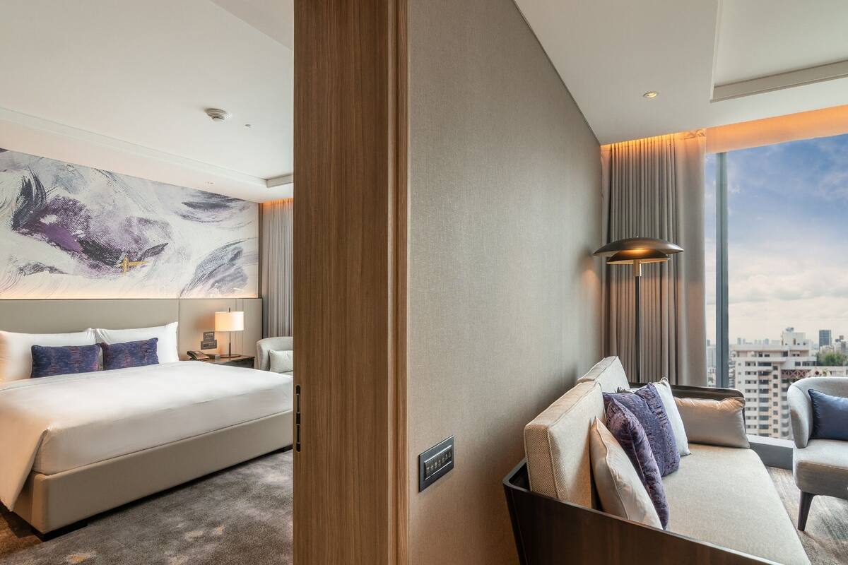 A modern hotel room with a large bed, a sofa, a desk, and a city view.