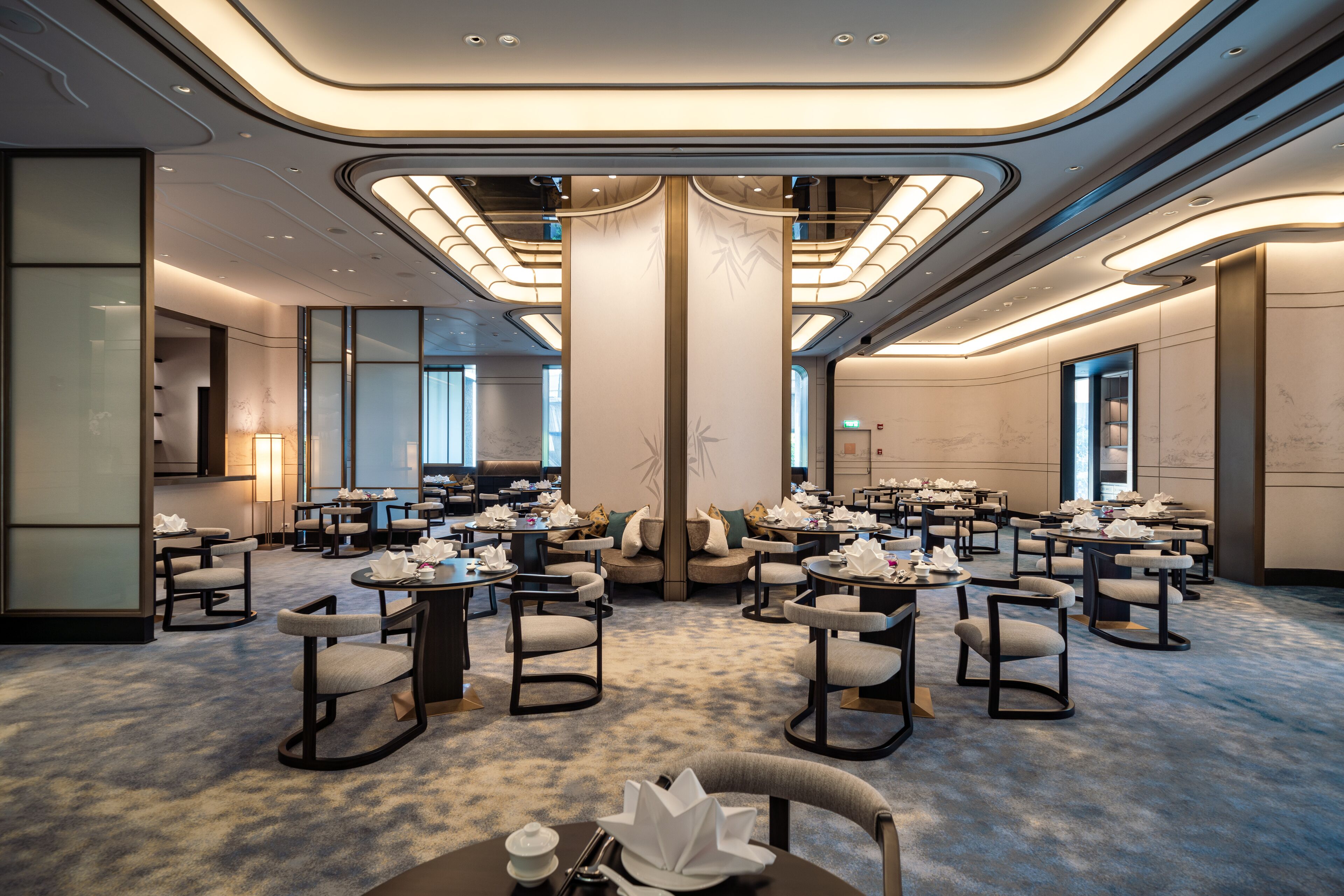 A modern hotel lobby with round tables and chairs, a central pillar, and a large window.