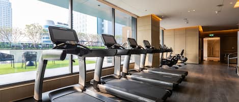 Una hilera de cintas de correr con pantallas digitales, suelo de madera y grandes ventanales con vistas a un paisaje urbano.