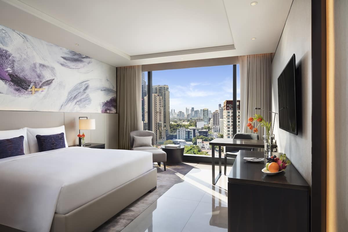A modern hotel room with a large bed, a desk, a chair, a TV, and a city view.