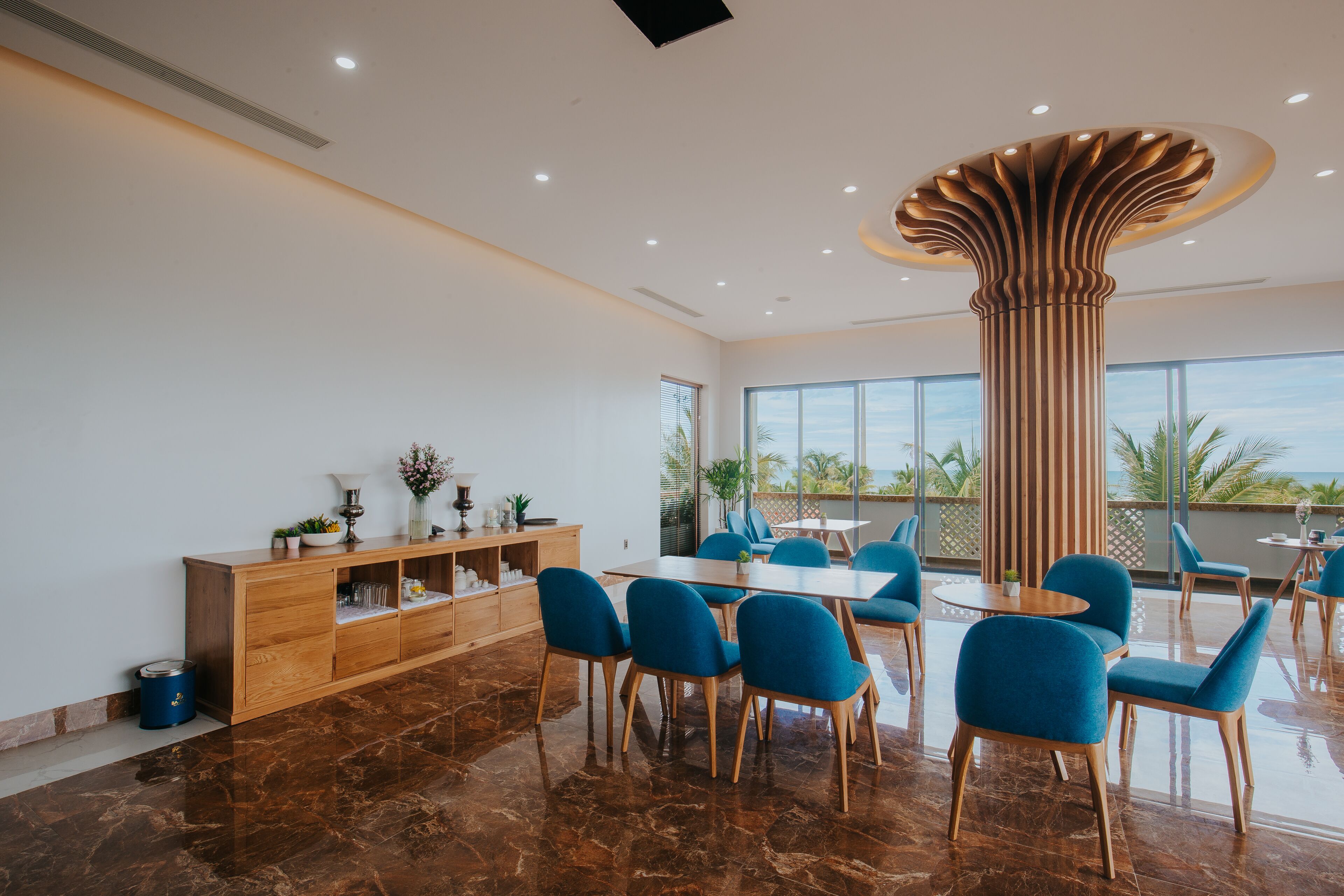 A modern dining area with a marble floor, wooden furniture, and large windows offering a view of palm trees.