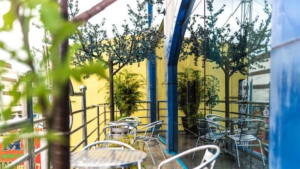 Un patio al aire libre con mesas y sillas de metal, rodeado de paredes amarillas y elementos estructurales azules.