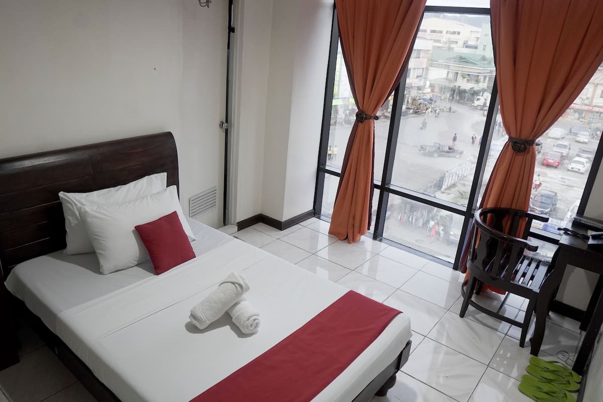 Habitación de hotel con cama, ventana con cortinas, vista a la calle y una silla cerca de la ventana.