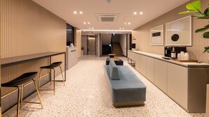 Een moderne hotellobby met een lange receptiebalie, twee barstoelen, een grijze bank en een keuken met een aanrecht en apparatuur.