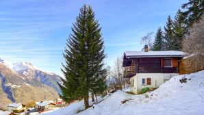 Una cabaña de dos pisos con un balcón, rodeada de nieve y montañas al fondo.