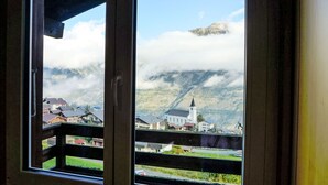 Vista a través de una ventana de un edificio con balcón, que da a un pueblo con casas, una iglesia y montañas parcialmente cubiertas de nubes.