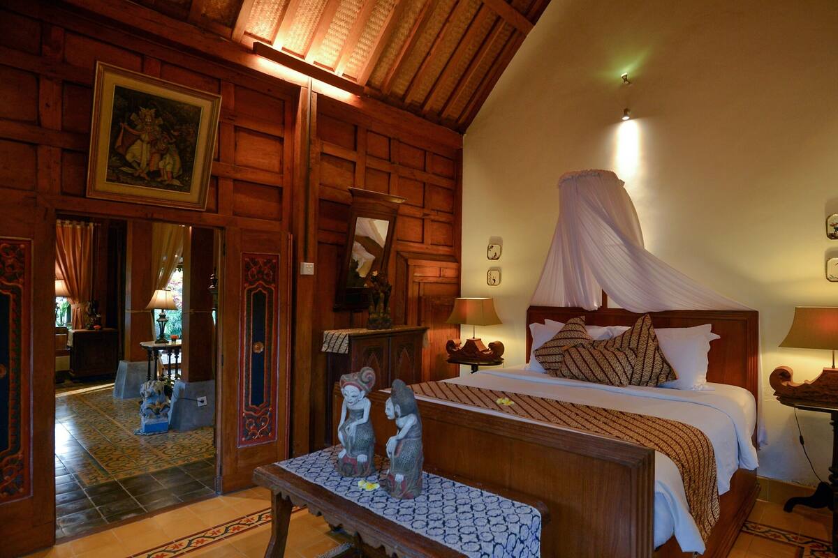 Una habitación tradicional con cama, lámparas de noche, un dosel y un banco decorativo.