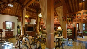 Amplia sala de estar con techos altos de madera, televisor, asientos cómodos y elementos decorativos como cortinas y arte tradicional.