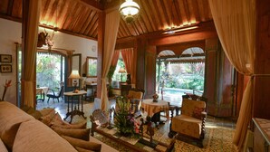 Amplia habitación con techo alto, vigas de madera y un gran ventanal con vistas a un jardín. Se puede ver un sofá, una mesa con sillas y una planta decorativa.