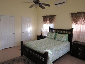 A bedroom with a bed, a ceiling fan, an air conditioner, a nightstand, and a window with curtains.