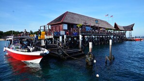 A wooden pier with a restaurant on stilts over water, a red and white boat docked at the pier, and flags in the background. - Borneo Divers Mabul Resort (Mabul Island)