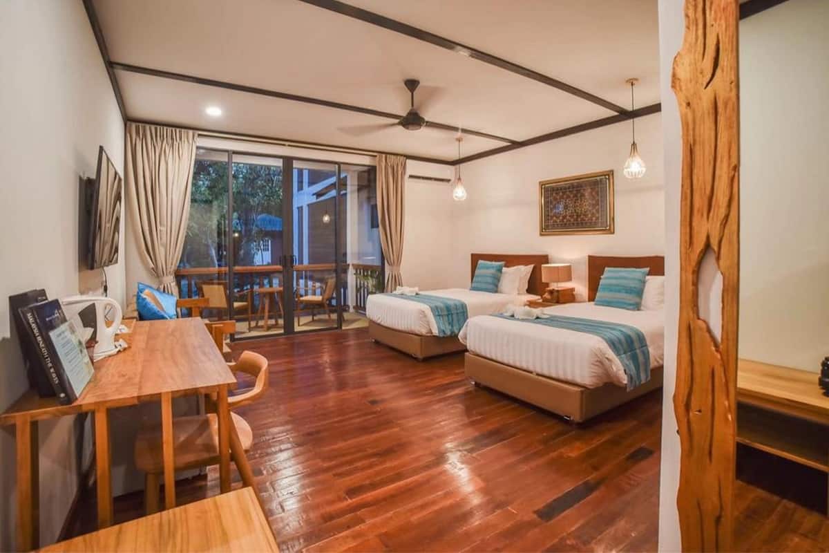 Une chambre d’hôtel avec deux lits, une table à manger en bois, un ventilateur de plafond et une grande fenêtre avec des rideaux.