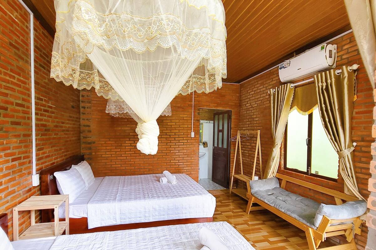 A room with two beds, a wooden bench, a hanging canopy, and a window with curtains.