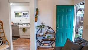 A kitchen with white cabinets and a stove, a teal door, a black metal shelf with plants, and a brown couch. - End of Summer Savings by the beach at Green Door! (San Diego)