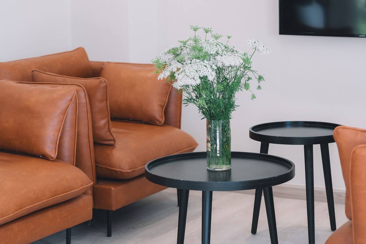 Una sala moderna con un sofá de cuero marrón, una mesa de centro redonda negra con un jarrón de flores blancas y un televisor negro montado en la pared.
