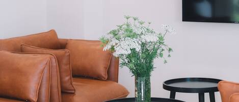 A modern living room with a brown leather sofa, a round black coffee table with a vase of white flowers, and a black TV mounted on the wall.