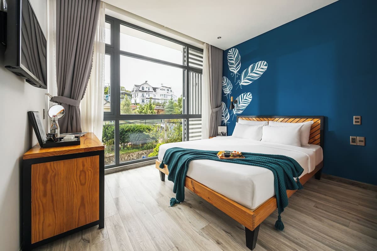 Un dormitorio con un ventanal grande, una cama con ropa de cama blanca, una mesita de noche de madera y una pared azul decorativa.