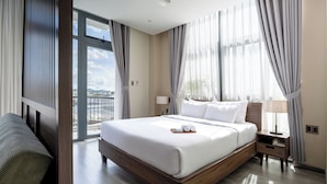 Una habitación de hotel moderna con una cama grande, un cabecero de madera, una mesita de noche con lámpara y vistas de la ciudad a través de amplios ventanales.