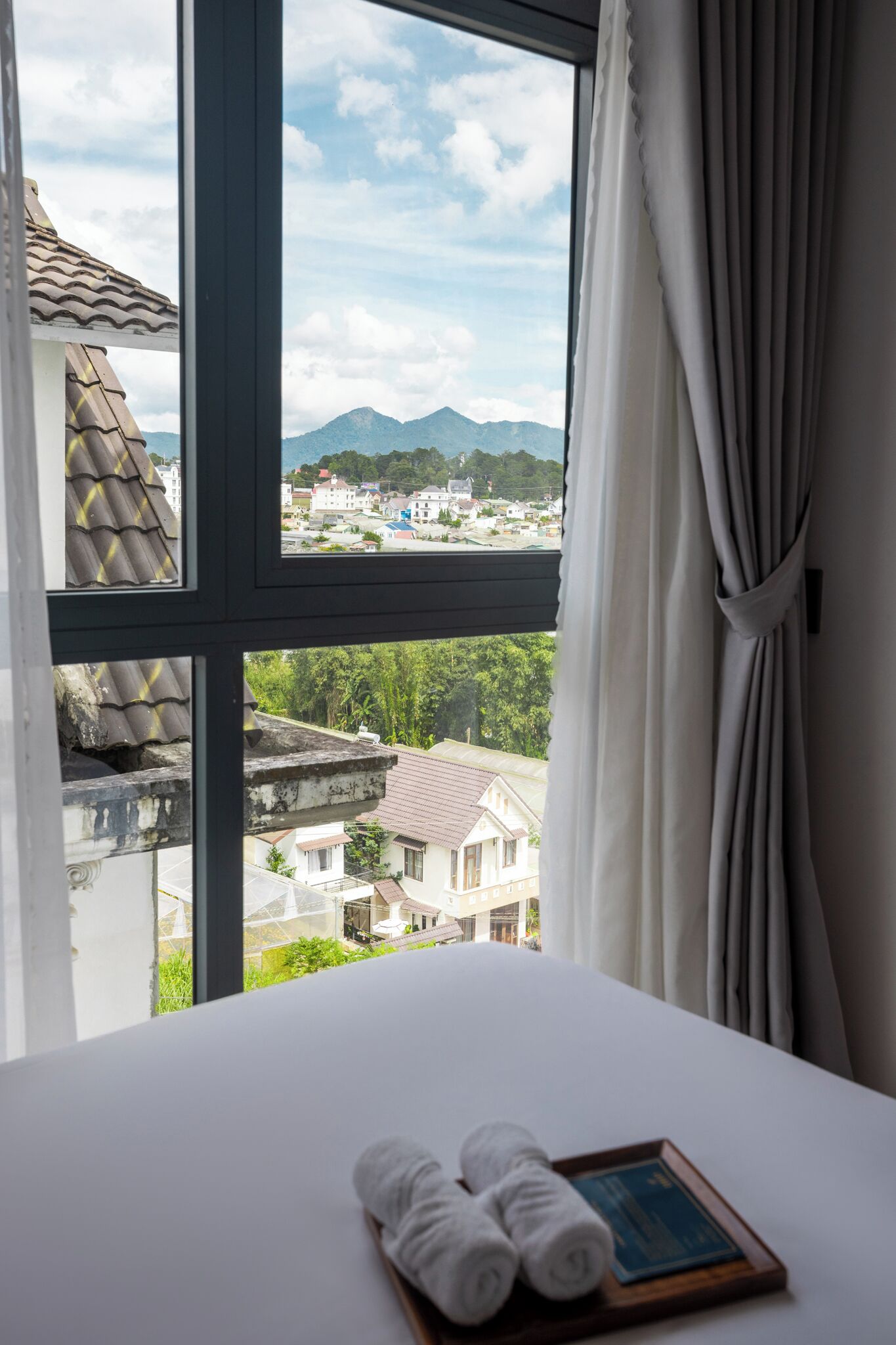 A hotel room with a large window, a neatly made bed, and a tray with towels and a book.