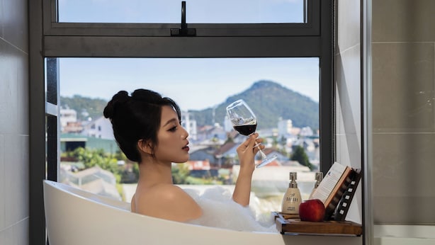 Una persona en una bañera con un libro y una botella de champú, sosteniendo una copa de vino, con vistas a edificios y montañas.