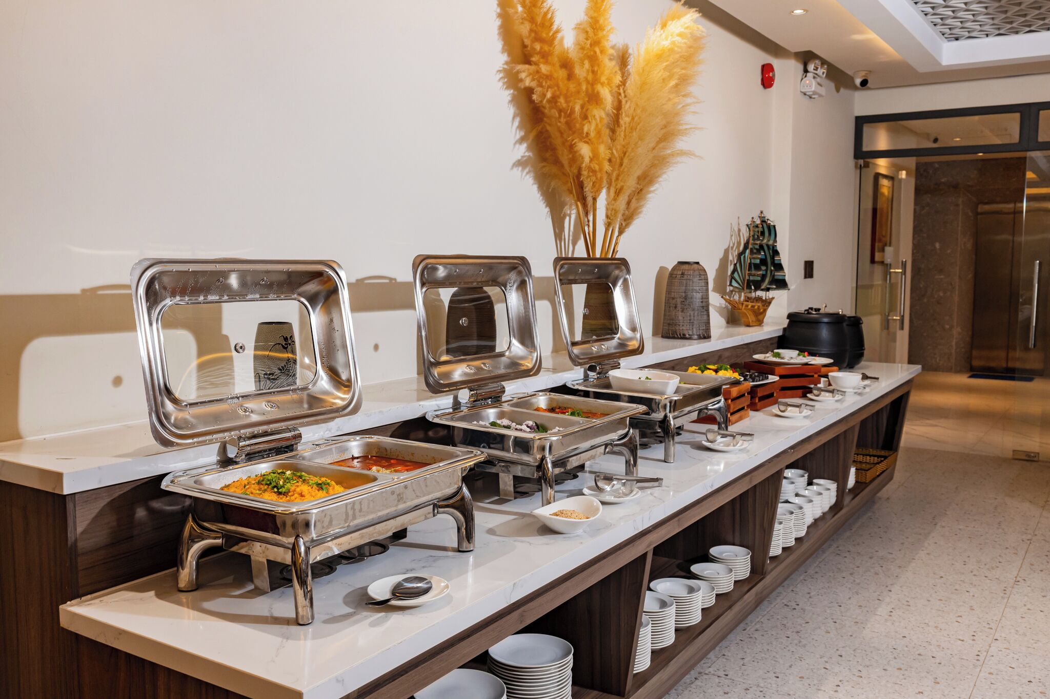 A buffet setup with multiple serving stations, a decorative vase with pampas grass, and a small ship model on the counter.